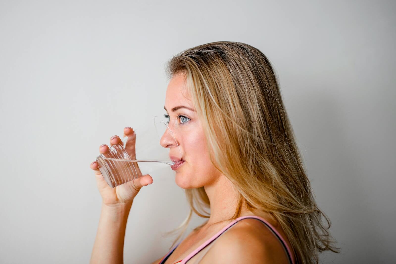 Mulher loira bebendo um copo de água com luz natural, representando a hidratação como medida de prevenção da cistite e das infecções urinárias