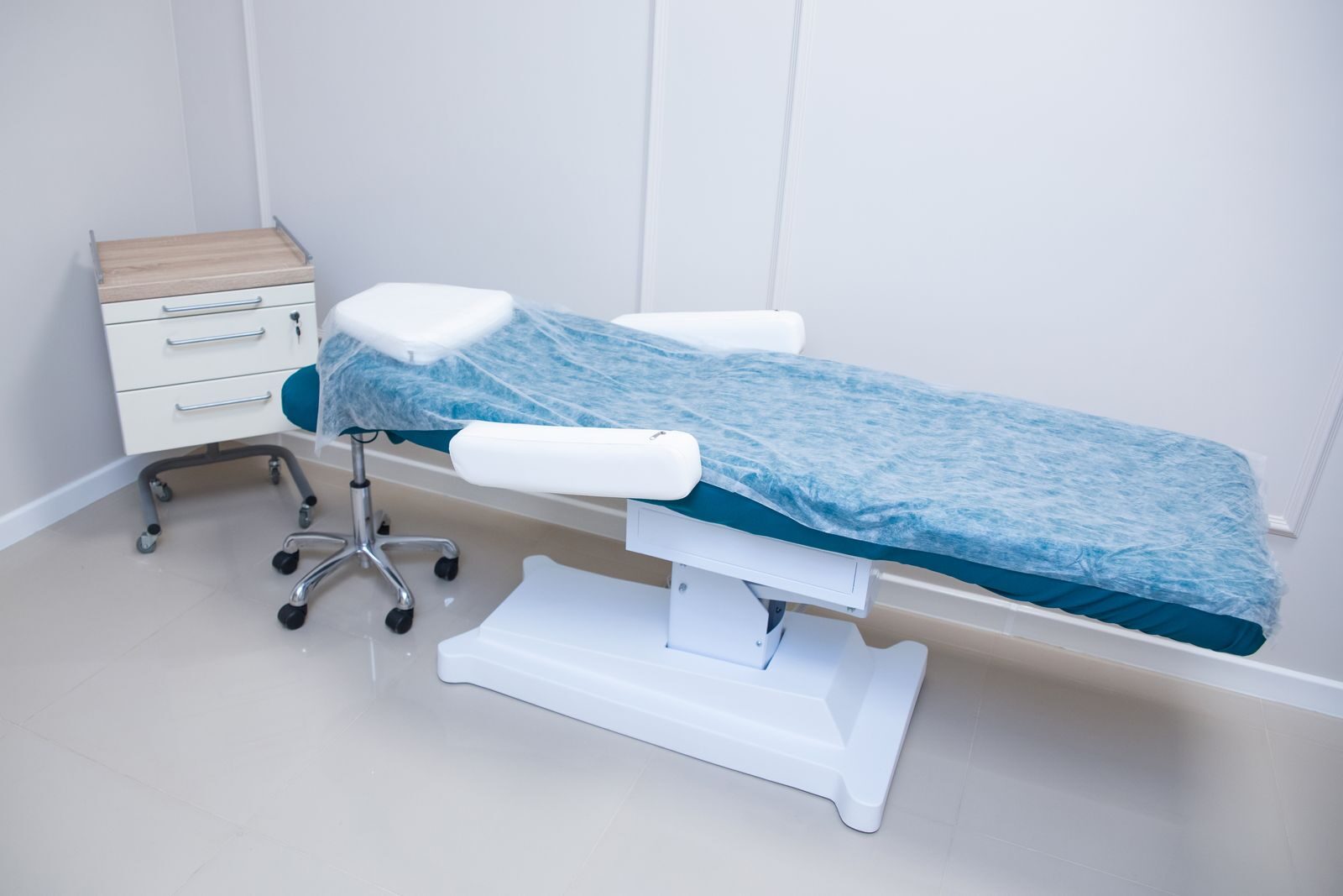 Sala de exame de uma clínica ginecológica, limpa e iluminada, com maca de procedimento na cor verde-azulada, mesa de atendimento e cadeiras, transmitindo ambiente tranquilo e profissional