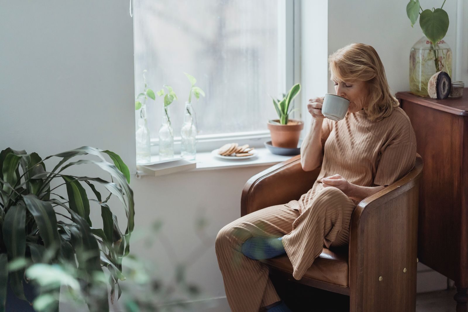 Mulher tranquila tomando uma xicara de cafe sentada em uma poltrona ao lado da janela, em uma sala iluminada e cheia de plantas, transmitindo bem-estar e conforto em casa