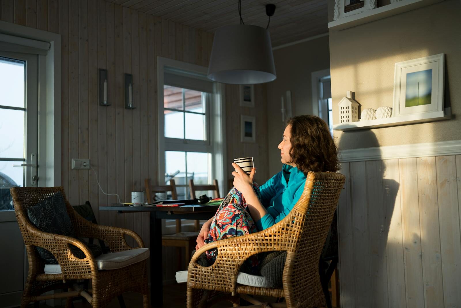 Mulher tranquila e sorridente relaxando em casa com uma xícara de café, sentada em uma poltrona perto da janela com luz natural