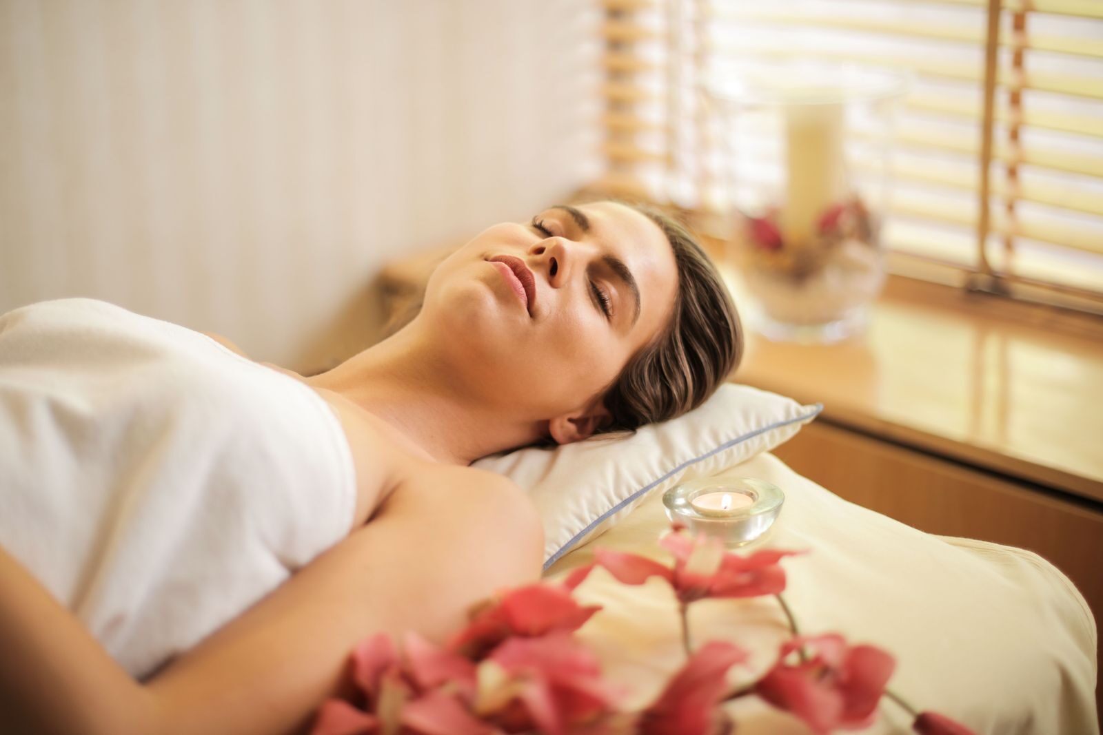 Mulher relaxada enrolada em toalha branca durante um dia de spa, cercada por orquideas e velas em um ambiente clean de bem-estar