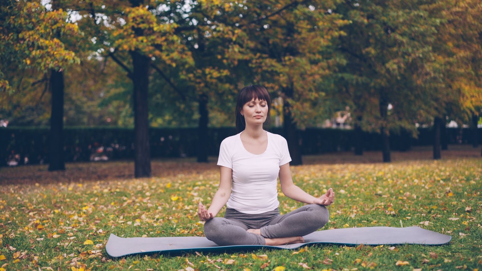 Mulher serena sentada em posição de meditação sobre um tapete de yoga em um parque arborizado no outono, com expressão tranquila e olhos fechados, transmitindo autocuidado e bem-estar.