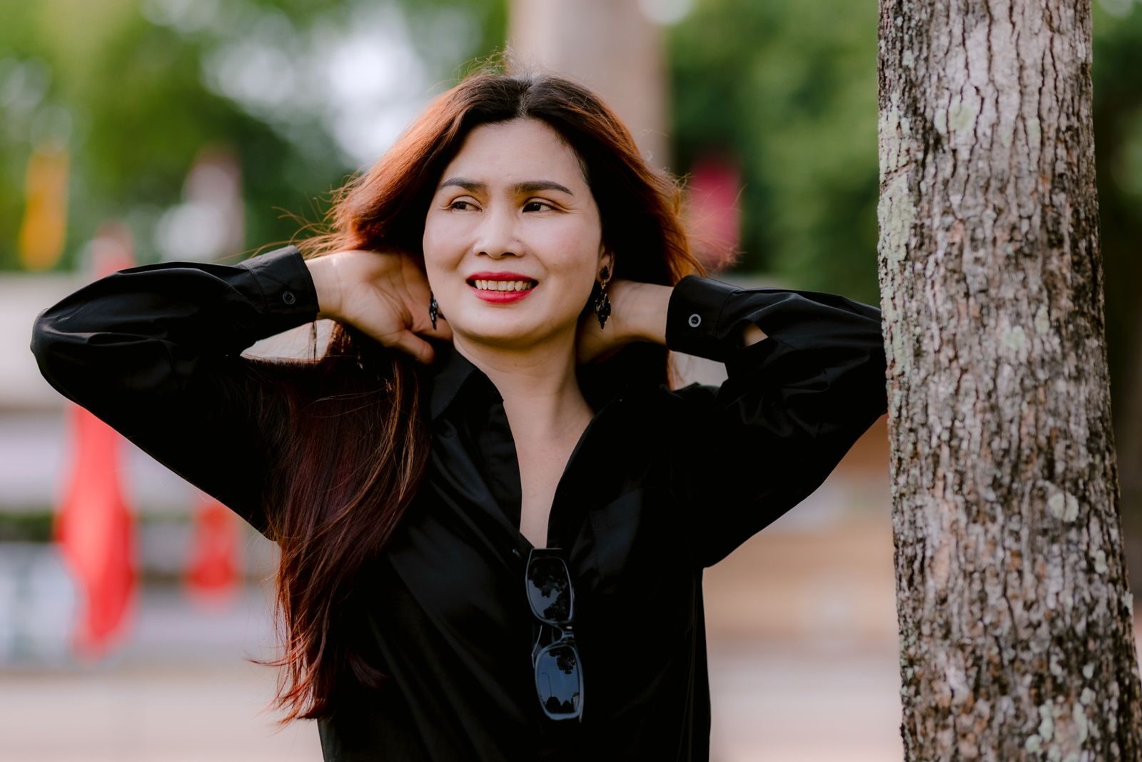 Mulher sorridente e relaxada ao ar livre, apoiada em uma árvore em um parque ensolarado, transmitindo bem-estar e saúde