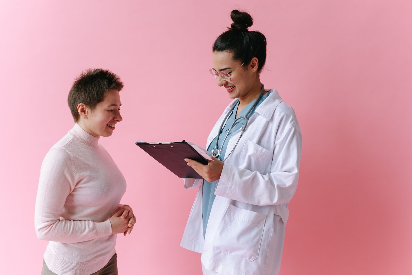 Médica sorridente de jaleco branco e estetoscópio orienta uma paciente durante consulta, segurando uma prancheta, em ambiente acolhedor de fundo rosa