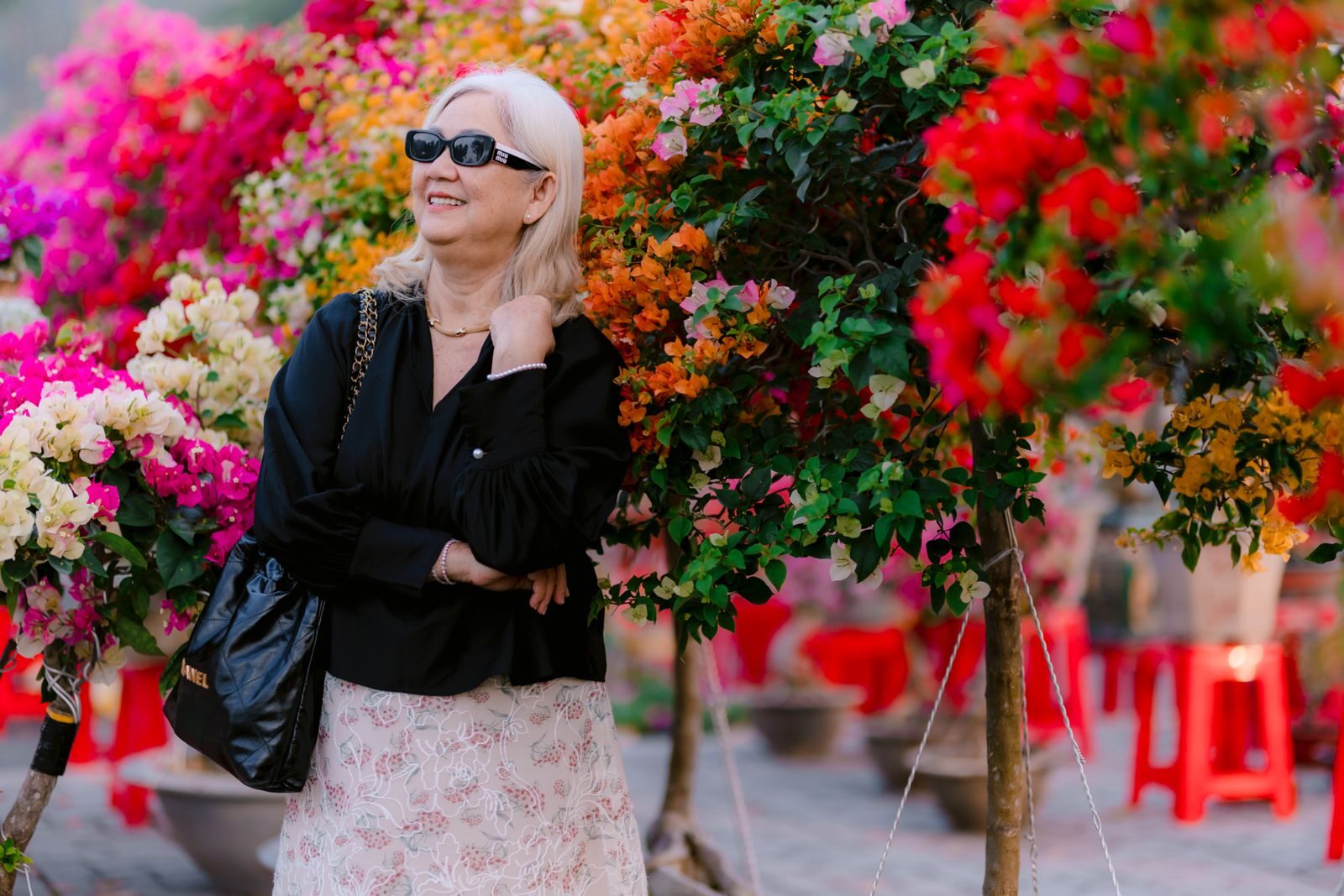 Mulher elegante e confiante, usando óculos de sol, sorrindo cercada por flores coloridas de buganvília em um jardim ensolarado, transmitindo bem-estar e leveza