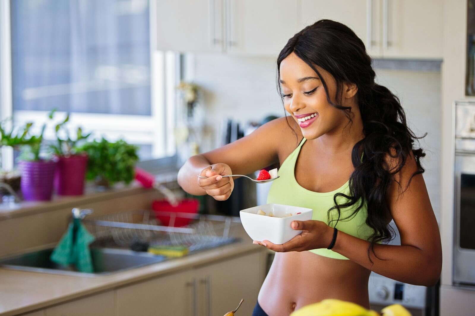 Mulher sorridente em uma cozinha aconchegante saboreando morangos frescos, transmitindo bem-estar, alimentação saudável e autocuidado no dia a dia.