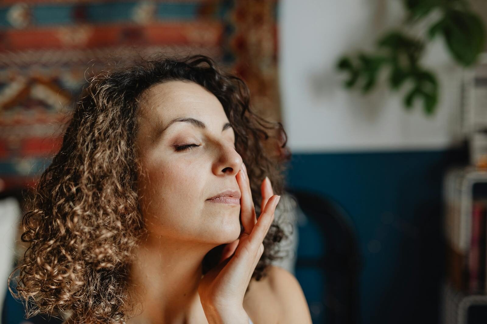 Mulher de cabelos cacheados em momento de autocuidado, com expressão serena e olhos fechados, tocando suavemente o rosto em um ambiente aconchegante e iluminado por luz natural
