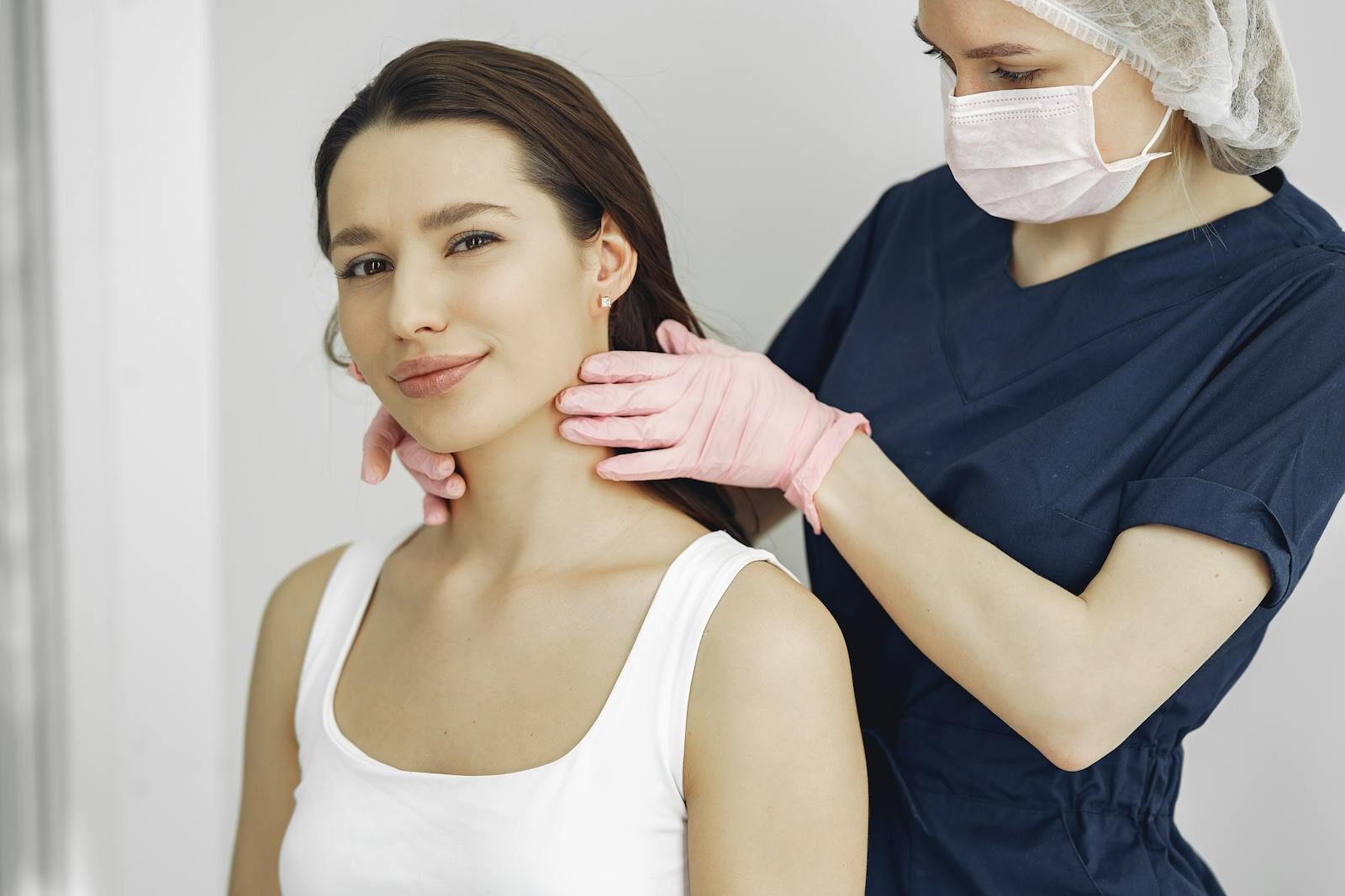Médica com luvas examinando a tireoide no pescoço de uma jovem paciente sorridente em consultório claro
