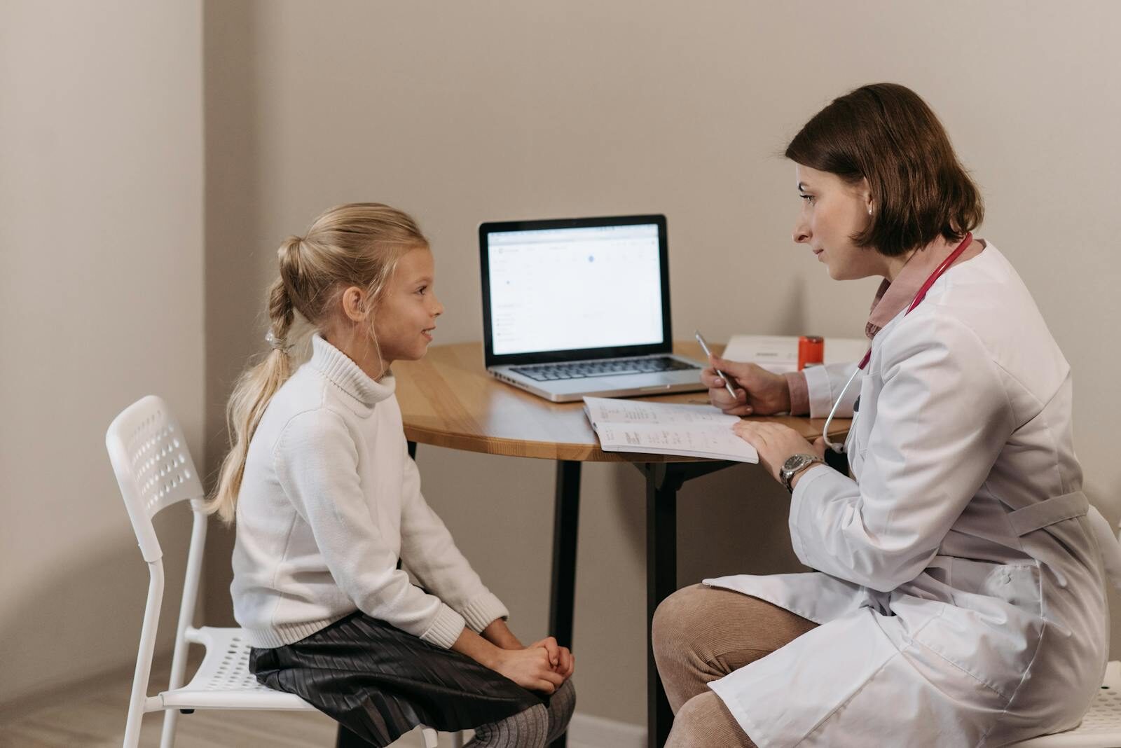 Médica de jaleco branco conversa de forma atenciosa com uma menina durante uma consulta em consultório claro e acolhedor
